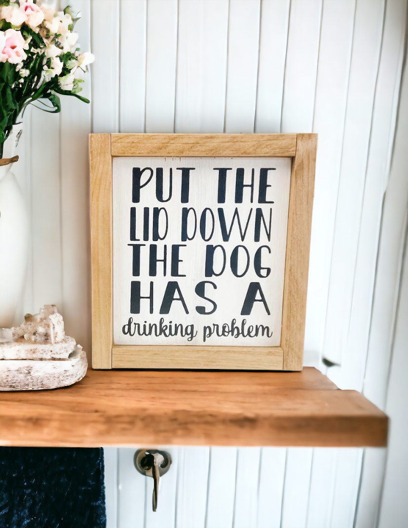 A 6.75" x 7.25" funny bathroom sign framed in stained golden oak wood. The sign features a white background with playful black text that reads, 'Put the lid down, the dog has a drinking problem.' This funny bathroom sign is the perfect dog sign to add humor and charm to your bathroom decor.