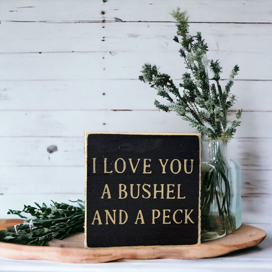 Hand-painted wooden block sign with black background and khaki text that reads 'I Love You a Bushel and Peck'
