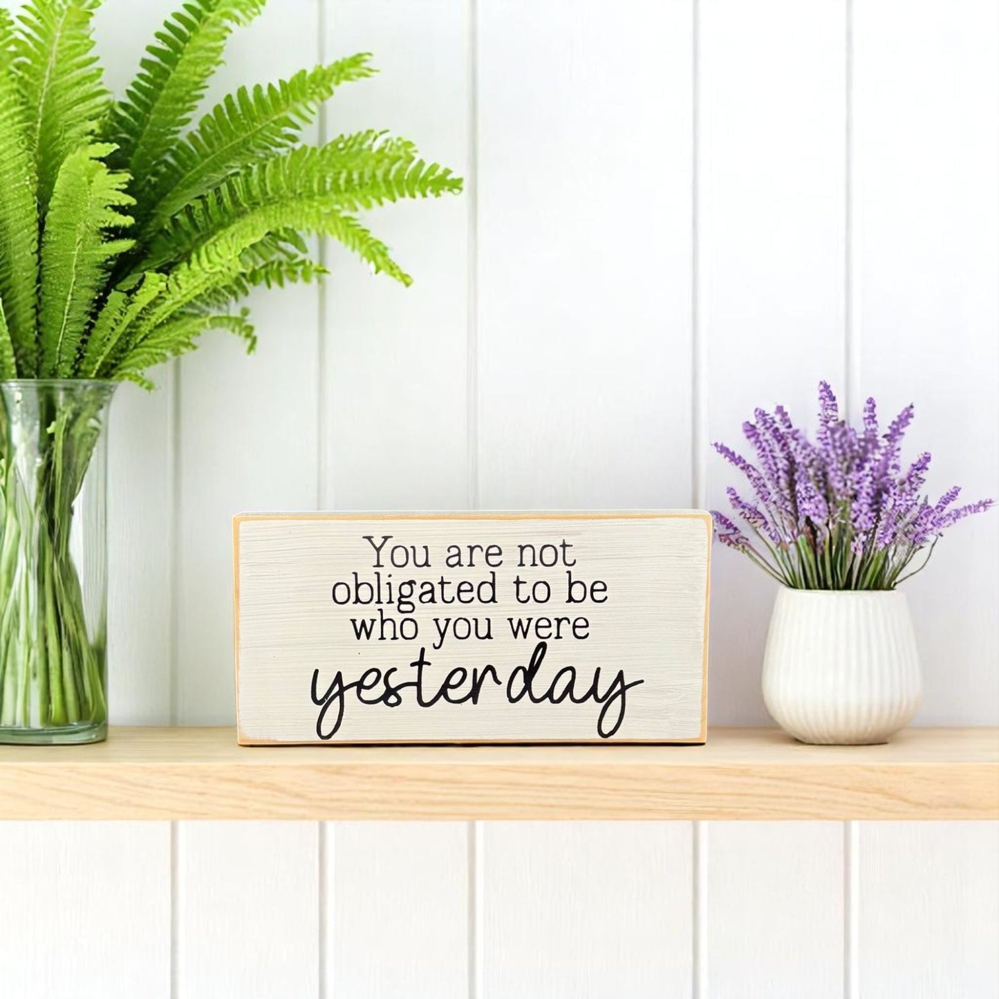 White wooden block sign with black text that reads, "You are not obligated to be who you were yesterday" – inspirational wood sign for shelf or desk.