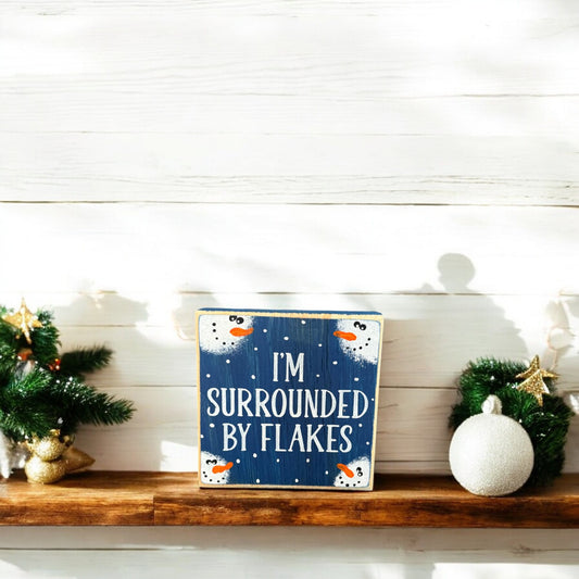 Handpainted wood sign with the phrase “I’m surrounded by flakes,” featuring snowman faces in each corner, perfect as Christmas mantel decor or a small coworker gift.