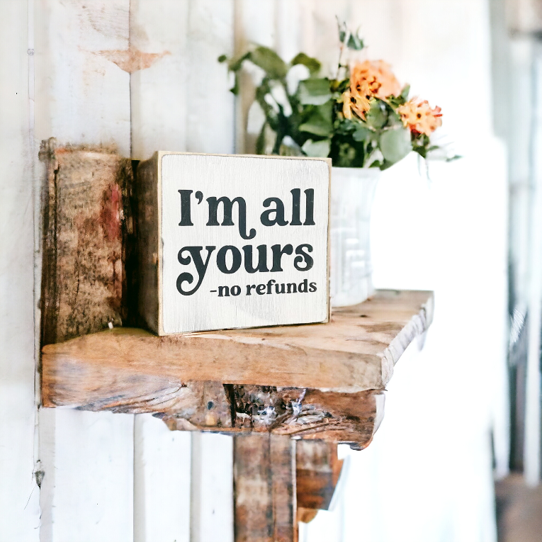 A 4.5" x 4.5" small wood sign with a white background and bold black text that reads, 'I'm all yours, no refunds.' This small wood sign is the perfect wood love sign, designed for couples who enjoy a touch of sarcasm and playful teasing, making it an ideal choice for a unique and light-hearted gift.