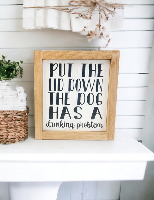 A 6.75" x 7.25" funny bathroom sign framed in stained golden oak wood. The sign features a white background with playful black text that reads, 'Put the lid down, the dog has a drinking problem.' This funny bathroom sign is the perfect dog sign to add humor and charm to your bathroom decor.