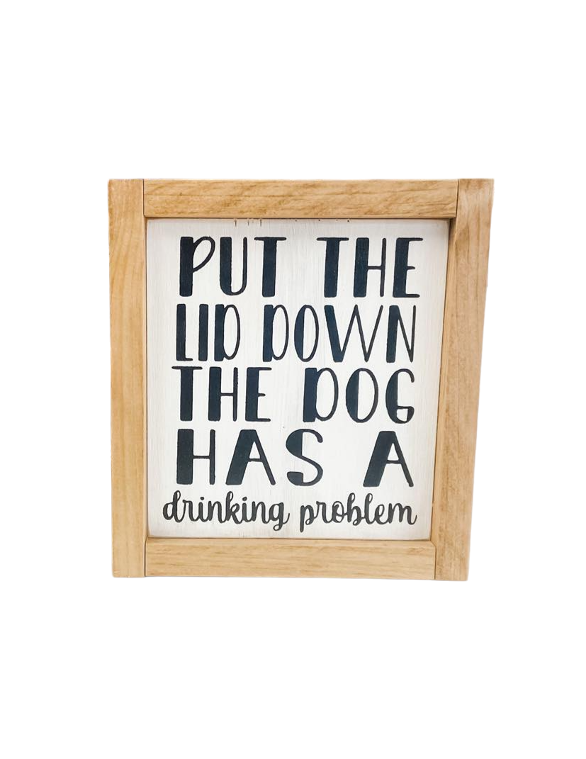 A 6.75" x 7.25" funny bathroom sign framed in stained golden oak wood. The sign features a white background with playful black text that reads, 'Put the lid down, the dog has a drinking problem.' This funny bathroom sign is the perfect dog sign to add humor and charm to your bathroom decor.