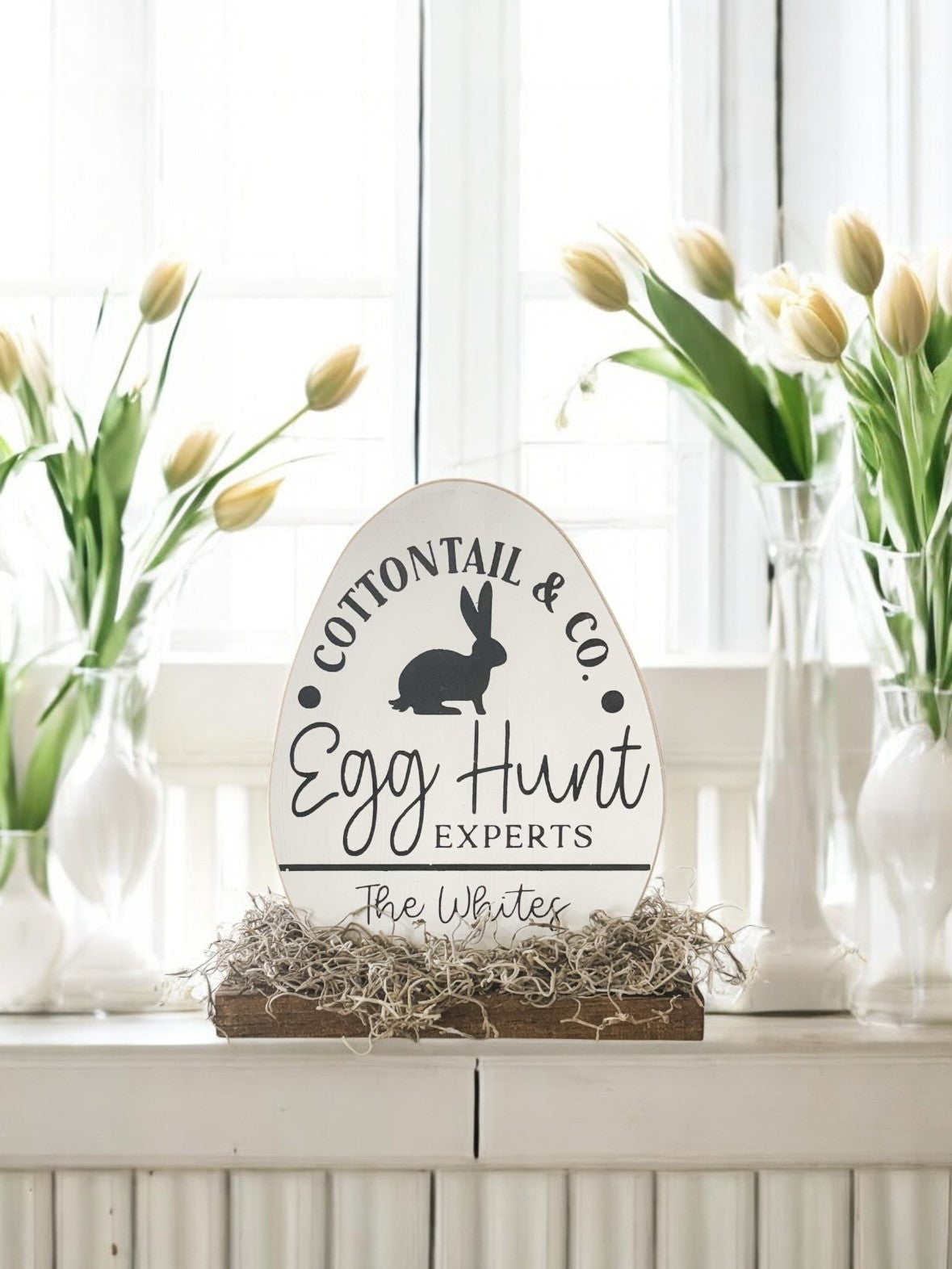 Personalized wooden Easter egg sign with "Cotton Tail Co. Egg Hunt Experts" in black text, featuring a family name, hand-painted white and attached to a stained wooden base, perfect for Easter décor.