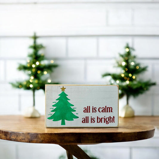 Handpainted wooden Christmas block sign that reads “All is Calm, All is Bright” in red text on a white background, featuring a cute green Christmas tree, ideal for Silent Night-themed holiday decor.