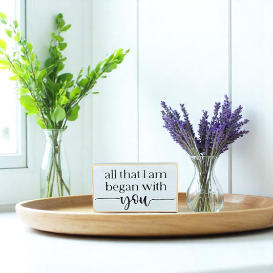 White wooden block sign with black text that reads “All That I Am Began With You” – meaningful gift for Mother’s Day.

