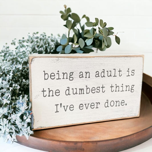 Funny desk sign with white background and gray text: 'Being an Adult Is the Dumbest Thing I've Done