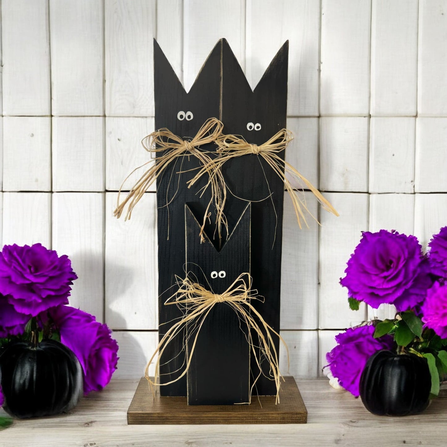 Handcrafted black cat decoration for Halloween, featuring three black cats with raffia bows on a stained wooden base. Ideal for rustic fall porch and mantel decor.