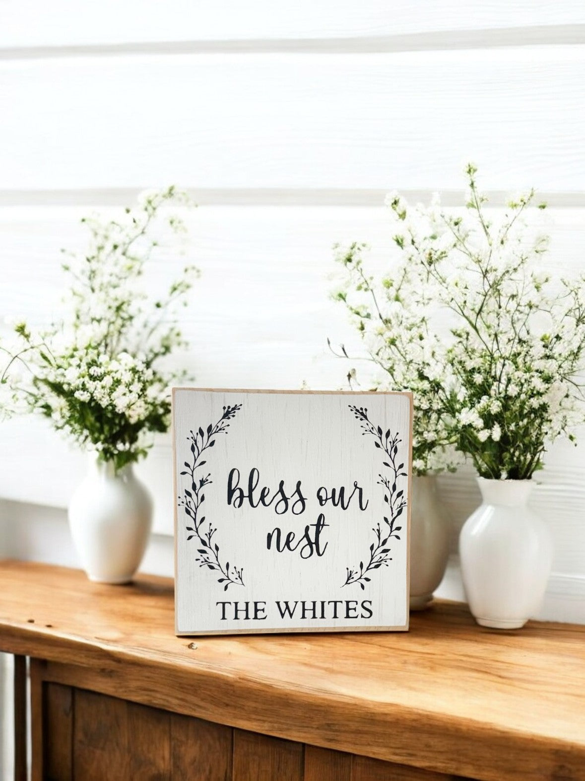  Personalized 5.5" x 5.5" wooden block sign with "Bless Our Nest" in black text, surrounded by a delicate spring floral wreath and a custom family name, hand-painted in a rustic modern farmhouse style.