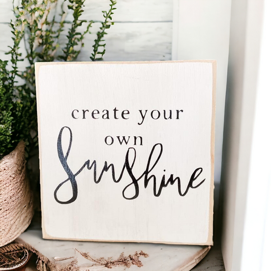 A 4.5" x 4.5" small wood block sign with a white background and bold black text that reads, 'create your own sunshine.' This inspirational wood sign is a perfect desk sign and tabletop decor, serving as a daily reminder to infuse positivity into your workspace.