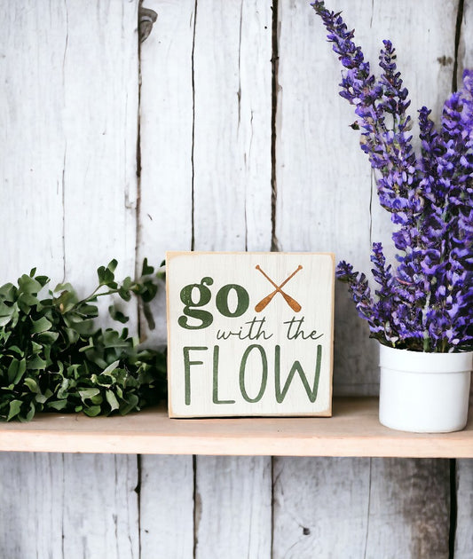 Image of wooden block sign with 'Go With The Flow' motto, adorned with paddles and earthy colors, perfect for camping decor.