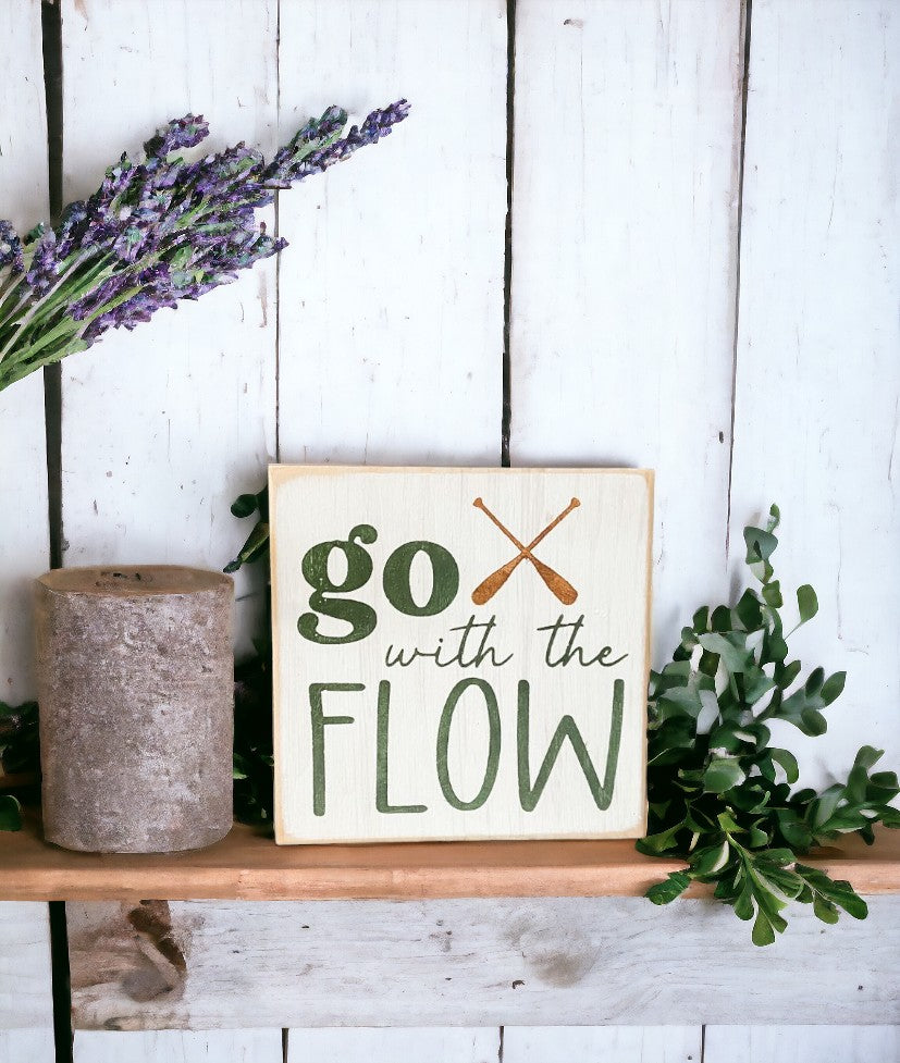 Image of wooden block sign with 'Go With The Flow' motto, adorned with paddles and earthy colors, perfect for camping decor.