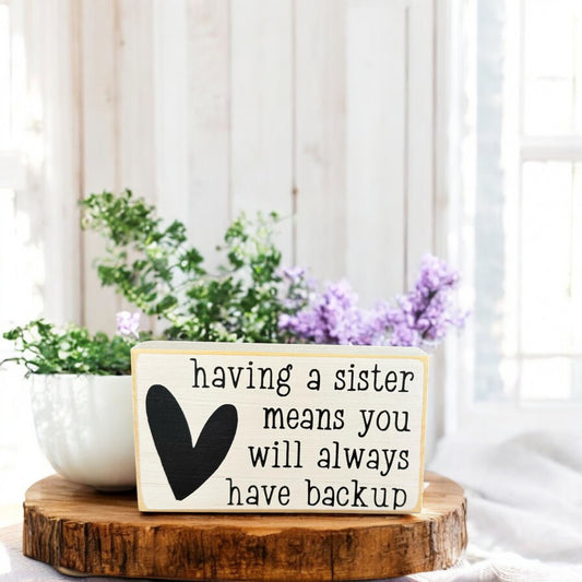 Handpainted white wood block sign with black text that reads, “Having a sister means you will always have back up,” perfect as a gift idea for a sister.