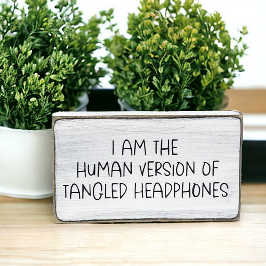 A 3.5" x 6" small wood sign featuring a white background with playful black text that reads, 'I am the human version of tangled headphones.' This small wood sign, designed with a touch of sarcasm, is a perfect and funny addition to your office decor.