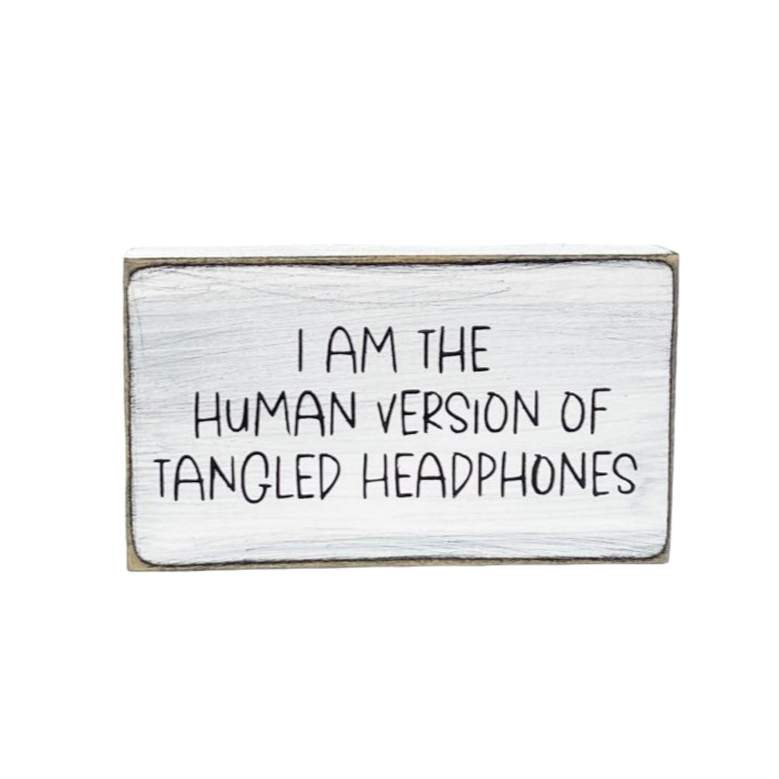 A 3.5" x 6" small wood sign featuring a white background with playful black text that reads, 'I am the human version of tangled headphones.' This small wood sign, designed with a touch of sarcasm, is a perfect and funny addition to your office decor.