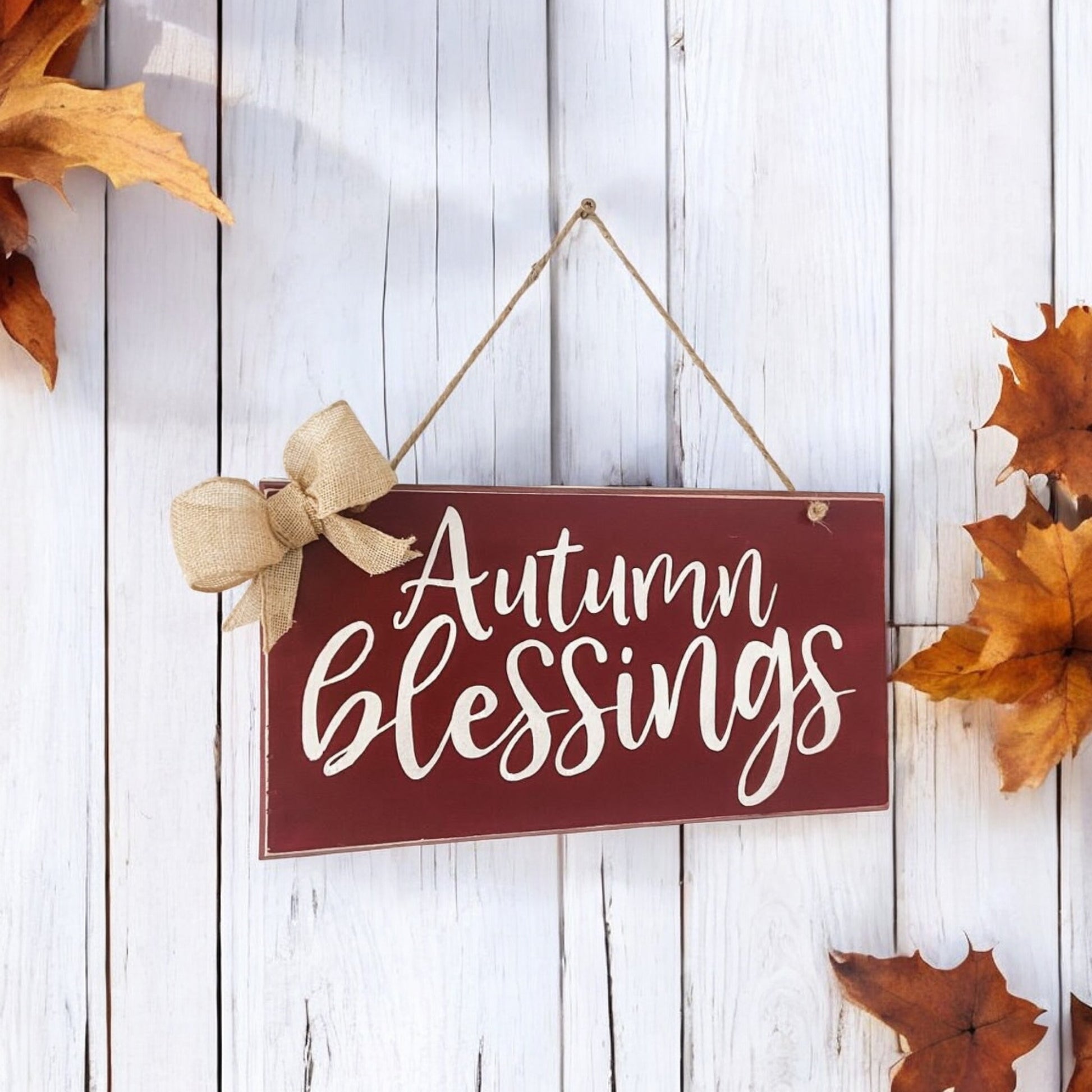 Handpainted 7.5" x 12" wooden fall sign with "Autumn Blessings" in maroon and white text, embellished with a burlap bow and a twine hanger for easy display.