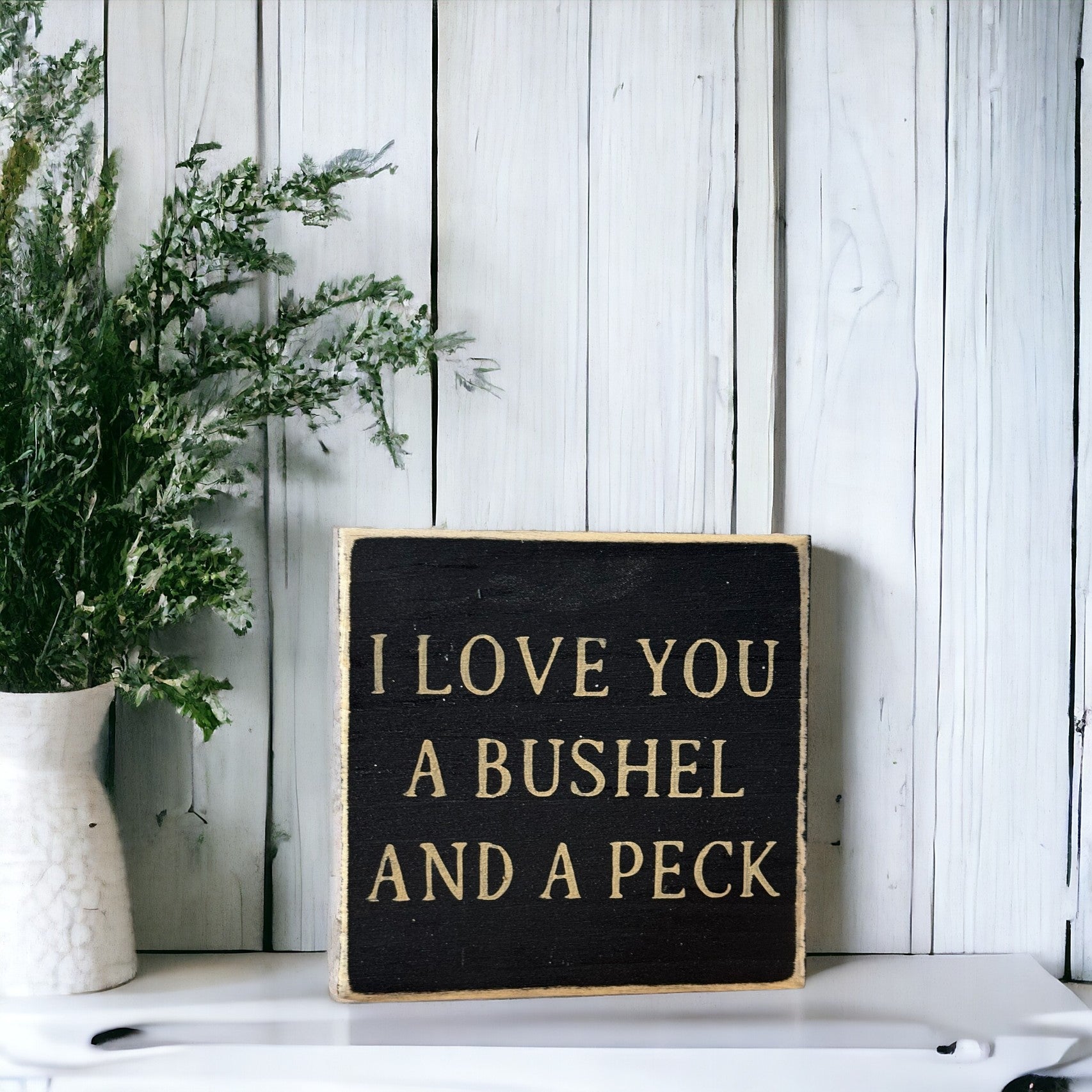Hand-painted wooden block sign with black background and khaki text that reads 'I Love You a Bushel and Peck'