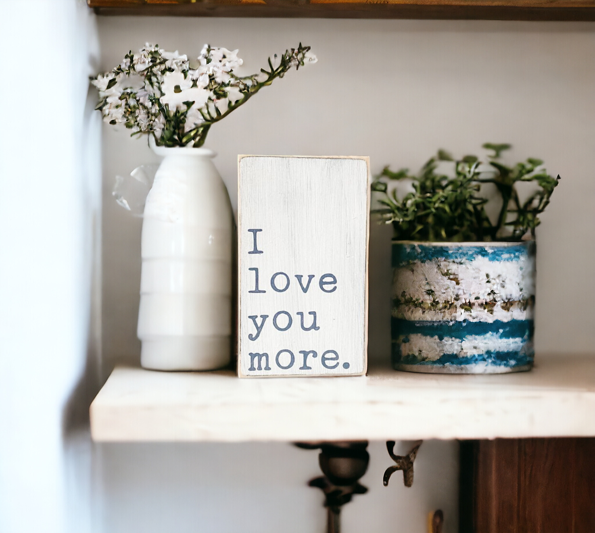 A 3.5" x 6" small wood sign with a white background and bluish gray text that reads, 'I love you more.' This versatile small wood sign adds a farmhouse-style touch to any space and makes a heartfelt gift for expressing your love.