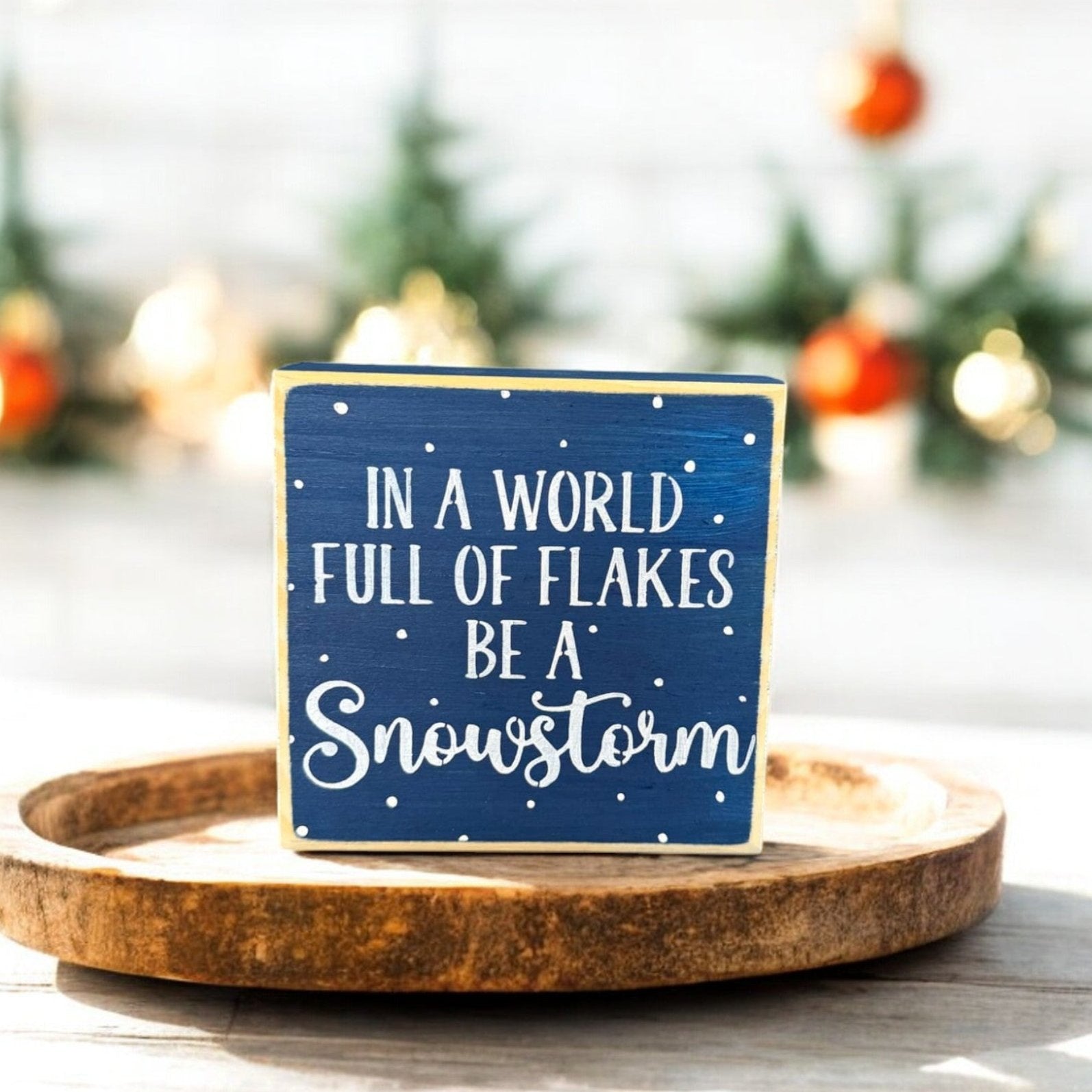 Handpainted wood sign with the inspirational winter quote “In a world full of flakes, be a snowstorm,” featuring navy blue background, white text, and surrounding snowflakes.