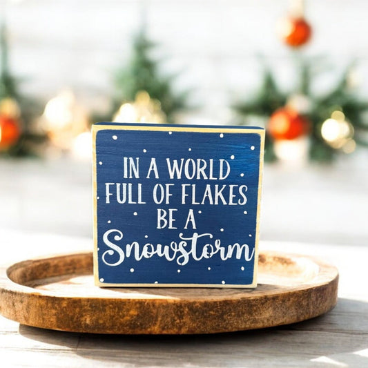 Handpainted wood sign with the inspirational winter quote “In a world full of flakes, be a snowstorm,” featuring navy blue background, white text, and surrounding snowflakes.