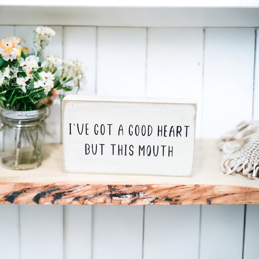 A 3.5" x 6" funny wood sign with a crisp white background and bold black text that reads, 'I've got a good heart but this mouth.' This freestanding sign is the perfect humorous gift, adding a touch of humor and sarcasm to your home or office decor as a funny office sign.