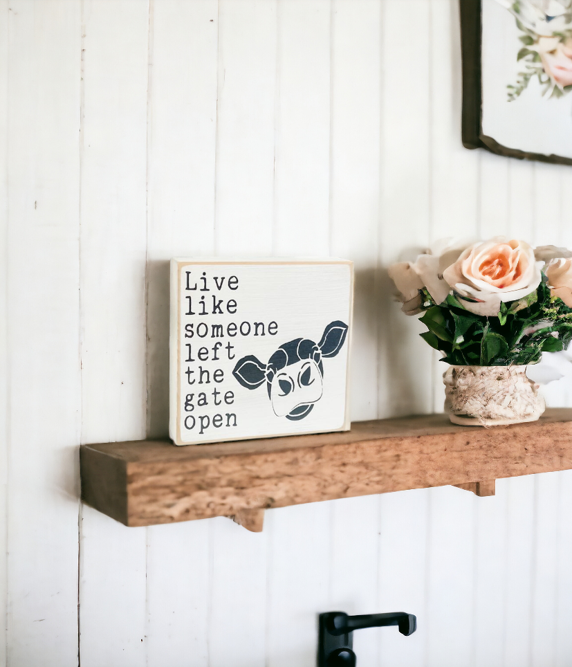 Small wood sign, 4.5" x 4.5", featuring a cute cow face and the phrase 'Live Like Someone Left The Gate Open' in bold black text on a white background. This charming cow sign stands on its own, making it a delightful addition to your decor.