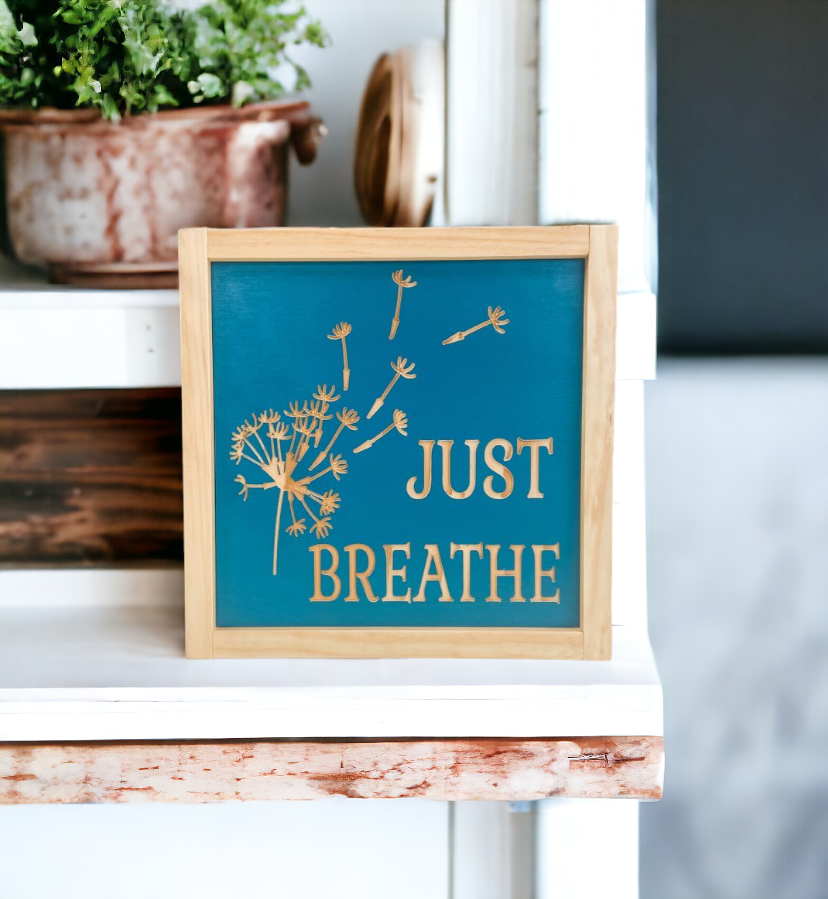 Carved wood sign, 9.5" x 9.5", with the words 'Just Breathe' and a dandelion image carved into natural wood, set against a soothing teal background. This sign adds a serene touch to your decor, offering a visual reminder to find moments of tranquility