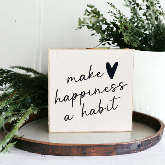 A 4.5" x 4.5" small wood sign with modern black text on a white background, displaying the inspirational happiness quote, 'Make Happiness A Habit.' This small wood sign is a charming addition to any space, serving as a reminder to prioritize joy and positivity