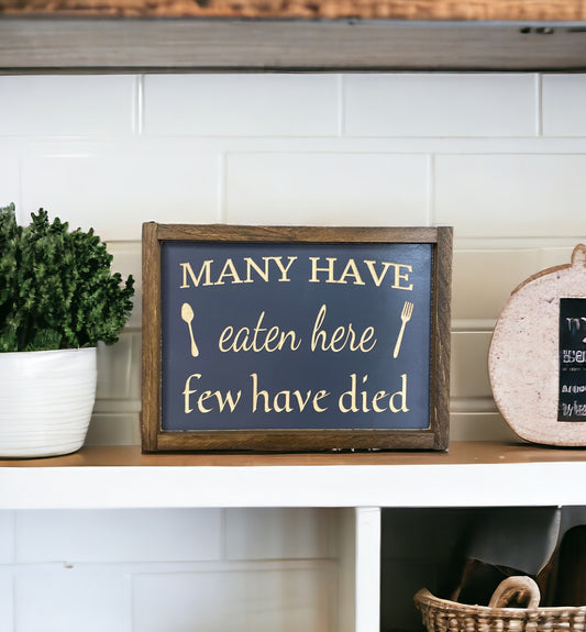 Handcrafted 'Many have eaten here, few have died' carved wood kitchen sign in bluish-gray with natural wood detailing