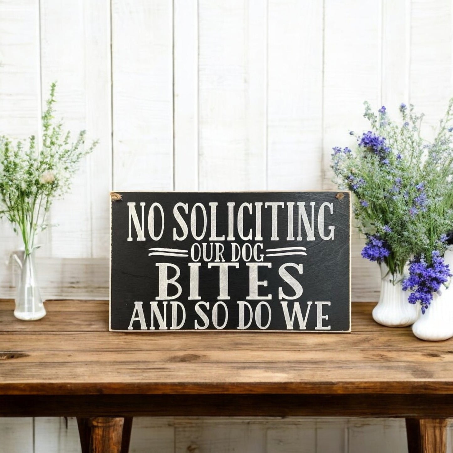 Wooden 'No Soliciting – Our Dog Bites and So Do We' sign, handpainted black with white text. Features a twine hanger for easy hanging. Funny beware of the dog sign for pet lovers.