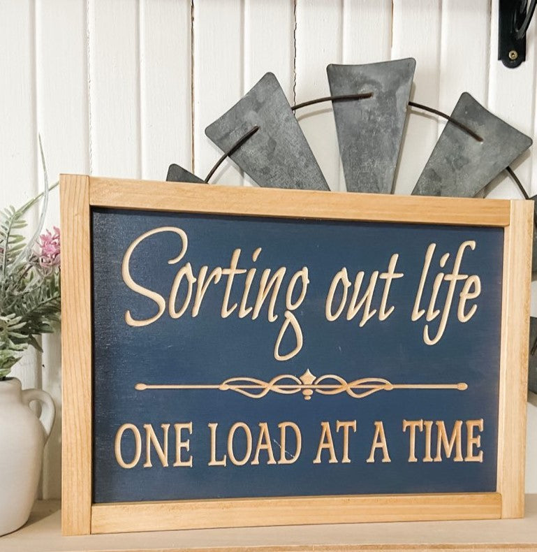 Carved wood sign for the laundry room, measuring 13.5" x 9.5", featuring natural wood text on a gray background that reads 'Sorting out life one load at a time.' This charming carved wood sign adds a touch of character and style to your laundry room decor