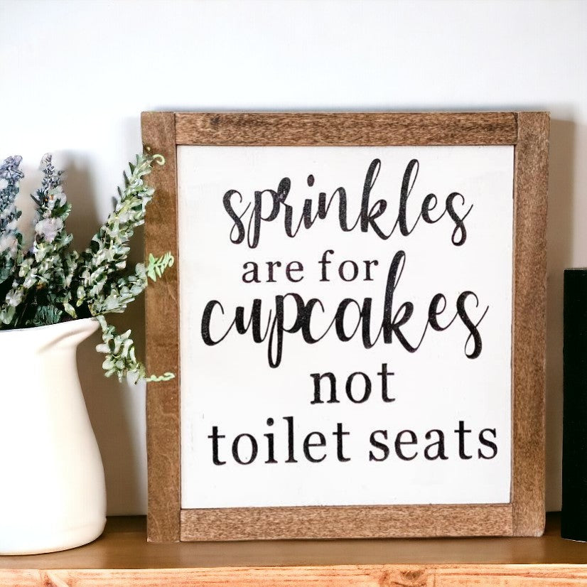 Framed wood sign with black text on white background, reading 'Sprinkles are for cupcakes not toilet seats', framed in dark stained wood
