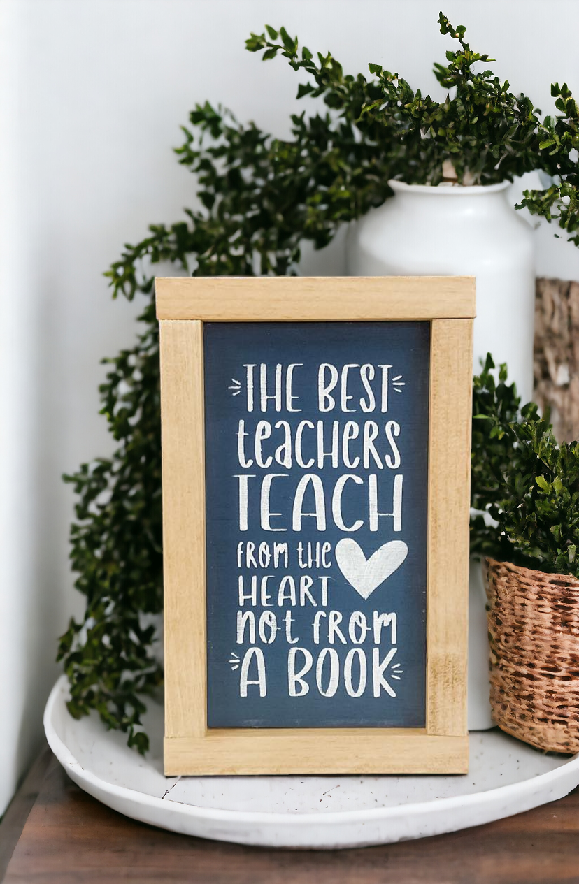 An 8" x 5" framed wood sign with a golden oak frame, featuring a bluish gray background and playful white text that reads, 'The best teachers teach from the heart not from a book.' This classroom sign is a thoughtful teacher gift for Christmas and an ideal choice for showcasing teacher appreciation quotes in educational spaces.