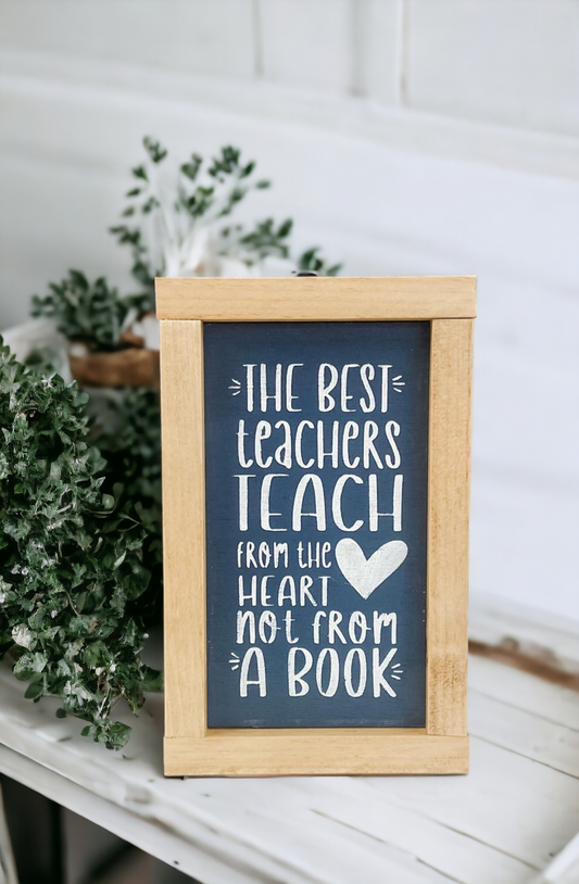 An 8" x 5" framed wood sign with a golden oak frame, featuring a bluish gray background and playful white text that reads, 'The best teachers teach from the heart not from a book.' This classroom sign is a thoughtful teacher gift for Christmas and an ideal choice for showcasing teacher appreciation quotes in educational spaces.