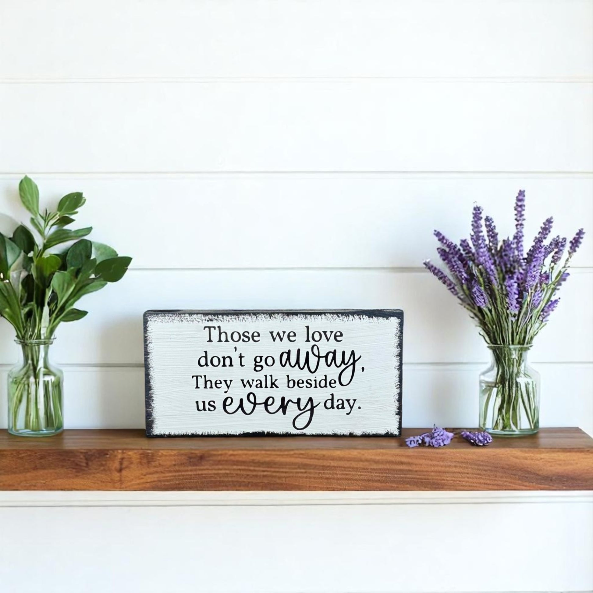 White handcrafted memorial wood sign with black text that reads, "Those We Love Don't Go Away. They Walk Beside Us Every Day."