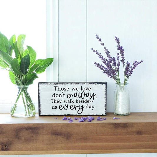 White handcrafted memorial wood sign with black text that reads, "Those We Love Don't Go Away. They Walk Beside Us Every Day."