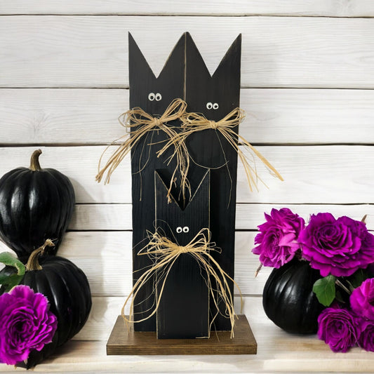 Handcrafted black cat decoration for Halloween, featuring three black cats with raffia bows on a stained wooden base. Ideal for rustic fall porch and mantel decor.