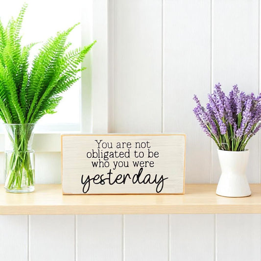 White wooden block sign with black text that reads, "You are not obligated to be who you were yesterday" – inspirational wood sign for shelf or desk.

