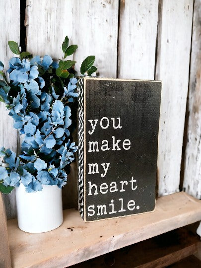 Hand-painted wood sign with white text on black background, featuring the phrase 'You Make My Heart Smile' surrounded by geometric design